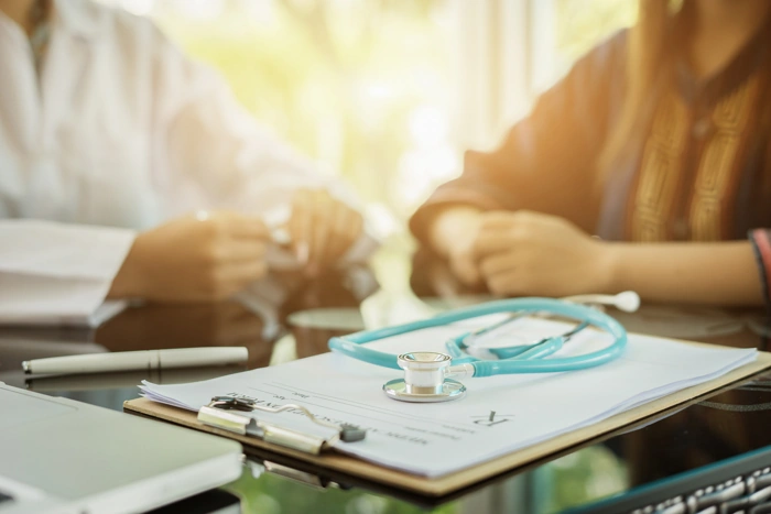 Médico conversa com paciente. Há um estetoscópio em cima da mesa, junto a uma caderneta. Explicando como cadastrar no sulamérica saúde boaconsulta.