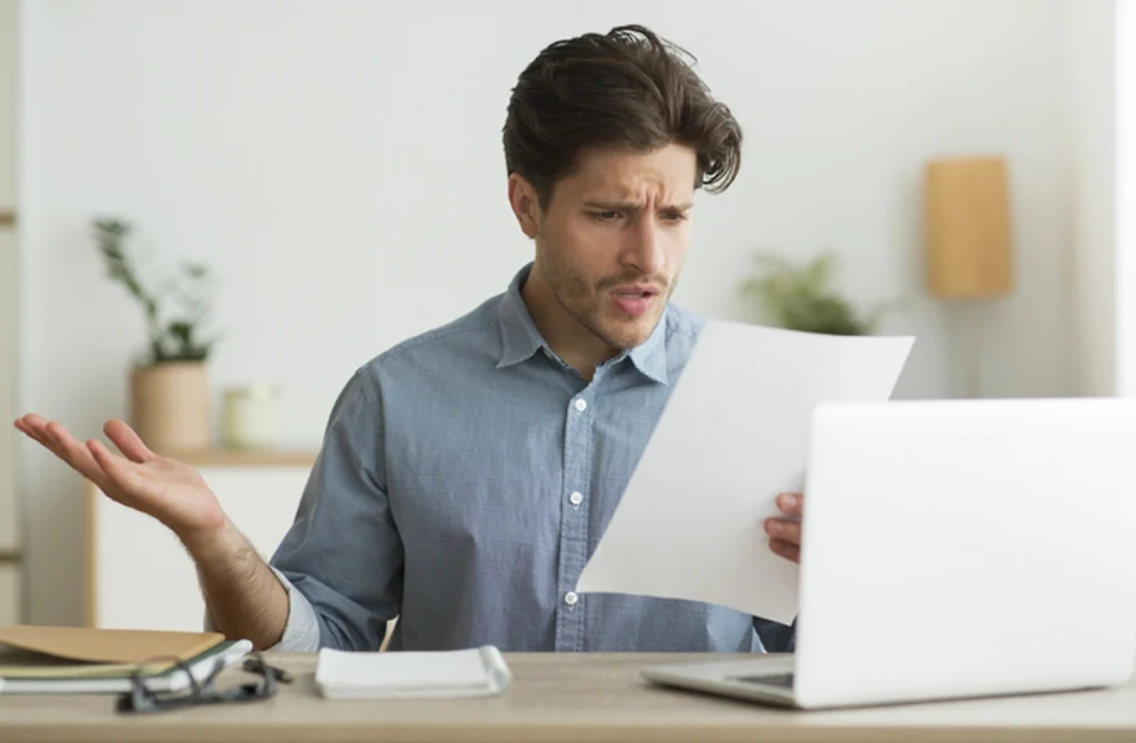 Homem analisa documento de plano de saúde e faz expressão de não entendimento. Há um notebook à sua frente. Explicando a lista de planos de saúde suspensos.